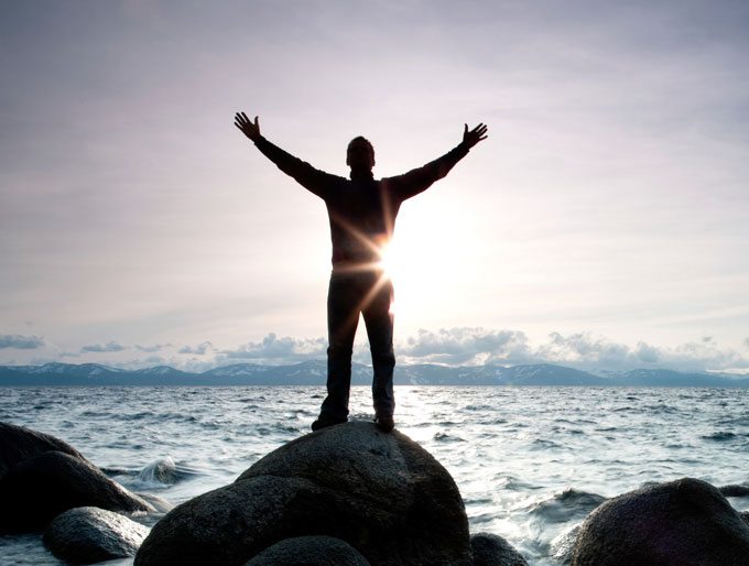 higher consciousness, man standing on rocks by ocean with outstretched arms.