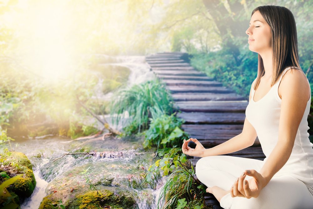 Benefits of meditation Close up side view of young woman in white meditating next to stream in forest.Girl sitting in wooden path next to water side.