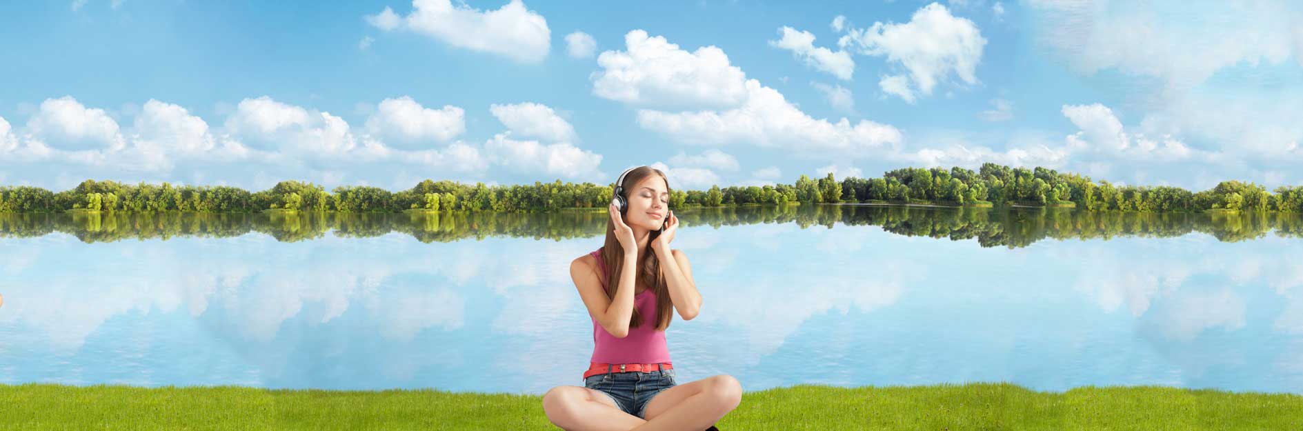 personal development a woman sitting cross legged listening to headphone on grass with a body of water behind her and clouds